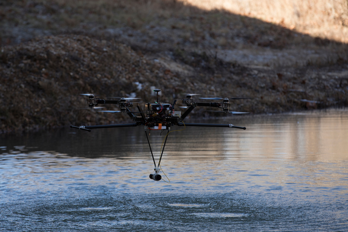 A multirotor drone hovers low over a calm lake, lowering a sensor into the water